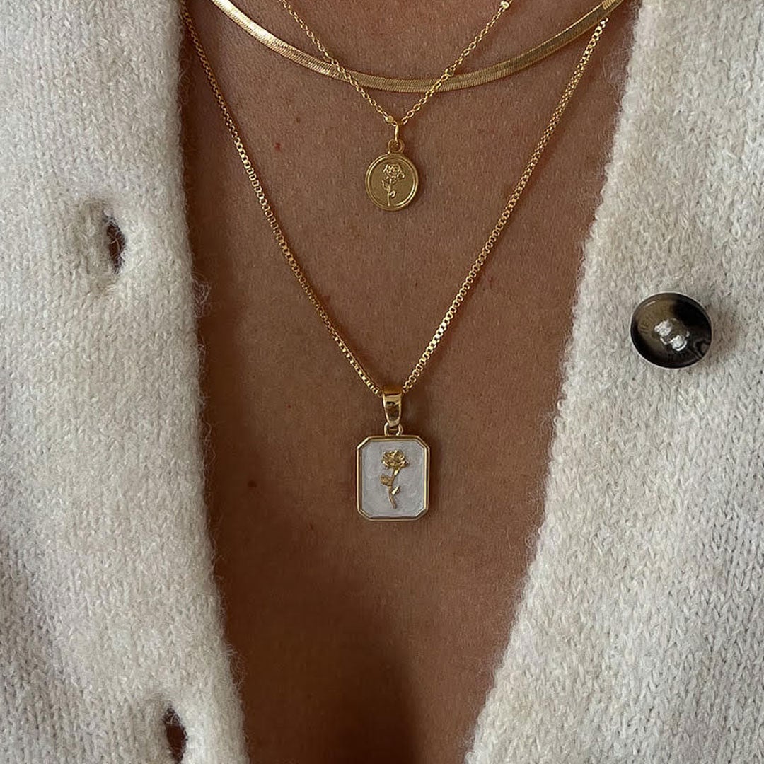 אדם הלובש סוודר בצבע שמנת עם שלוש שרשראות זהב בשכבות, שכל אחת מהן מעוטרת בתליון שונה, כולל מטבע עגול ועיצוב פרחוני מלבני.