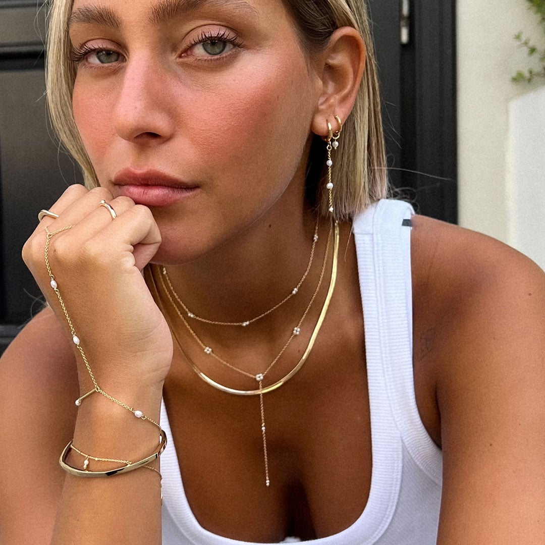 אישה לבושה בגופייה לבנה עומדת בתנוחה מהורהרת, עונדת שרשראות זהב בשכבות, צמידים, טבעות ועגילים עם נגיעות של פנינים.